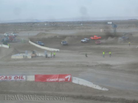 1990 Ford Bronco - Off-road race trucks crest dirt jumps on a rough course lined with Lucas Oil and Kumho Tires banners.