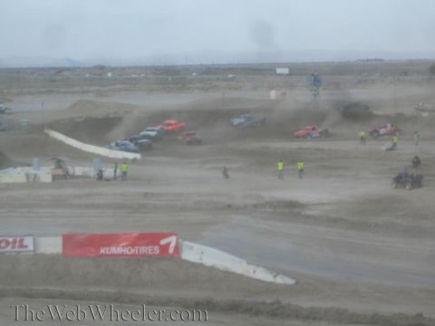 1990 Ford Bronco - Off-road race trucks run a dirt jump course as yellow-vested workers stand trackside.