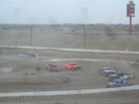 1990 Ford Bronco - Pink and orange off-road race trucks corner on a dusty dirt course near a red billboard.