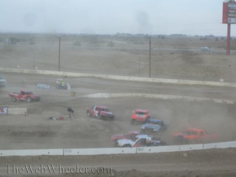 1990 Ford Bronco - Off-road race trucks crowd a turn on a dusty dirt course, kicking up clouds of dust.