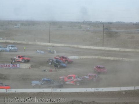 1990 Ford Bronco - tangle of off-road race trucks jostles in heavy dust on a desert dirt course with a Lucas Oil banner.