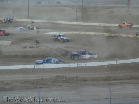 1990 Ford Bronco - Off-road race trucks battle through dust on a banked desert dirt course with Fabtech and Lucas Oil signage.