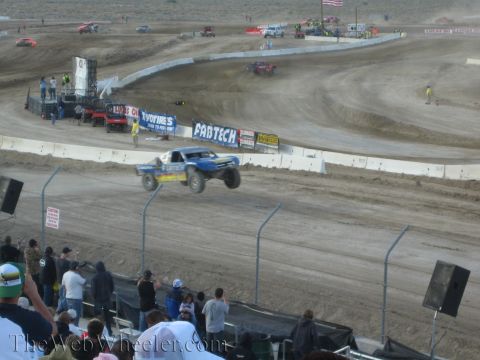 1990 Ford Bronco - blue off-road race truck catches air over a dirt jump as spectators watch beneath a Fabtech banner.