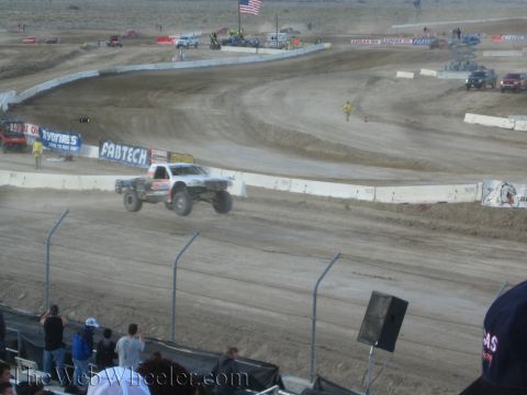 1990 Ford Bronco - off-road race truck launches off a dirt jump on a desert short-course track beneath a US flag.