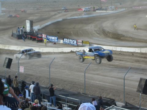 1990 Ford Bronco - Two off-road short-course race trucks throw up dust on a banked dirt track lined with sponsor banners as spectators watch.