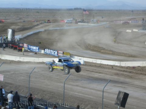 1990 Ford Bronco - blue and yellow off-road race truck slides through a dusty corner of a desert dirt track behind a chain-link fence.