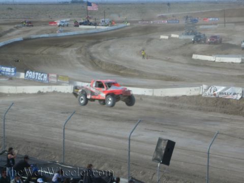 1990 Ford Bronco - red off-road race truck catches air over a jump on a dusty desert short-course track lined with sponsor banners.