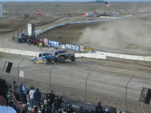 1990 Ford Bronco - blue off-road short-course race truck kicks up heavy dust on a banked dirt track as spectators watch from the stands.