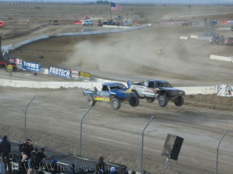 1990 Ford Bronco - blue and a black off-road race truck battle side by side through a dusty corner of a desert short-course track.