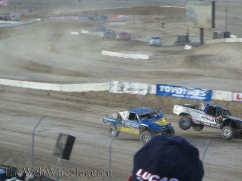 1990 Ford Bronco - Blue and white off-road race trucks race close together on a banked dirt course past Toyo Tires and Lucas Oil banners.