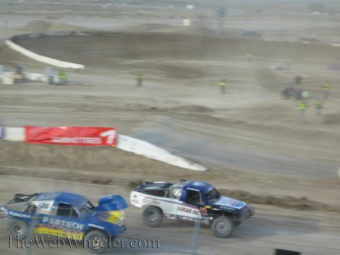 1990 Ford Bronco - Two off-road race trucks race along a dusty desert short-course track past a red banner and distant track workers.