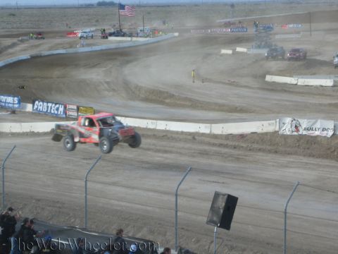 1990 Ford Bronco - red off-road race truck races along a dusty desert short-course track lined with sponsor banners and a chain-link fence.