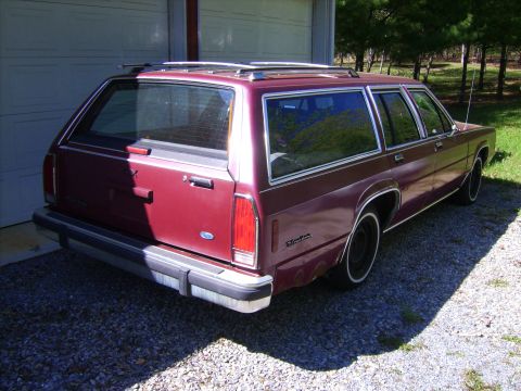 1982 Ford LTD Crown Victoria - Driver's-side rear three-quarter view of a maroon Ford full-size station wagon parked on gravel in front of a white garage door, with faded…
