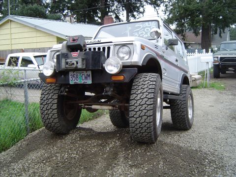 1989 Ford Bronco - Silver Suzuki Samurai equipped with a winch and large off-road tires is parked on a gravel incline. A second vehicle is visible in the back…