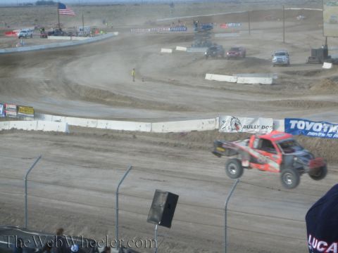 1990 Ford Bronco - red off-road race truck launches over a berm on a dusty desert short-course track past Toyo Tires and Bully Dog banners.