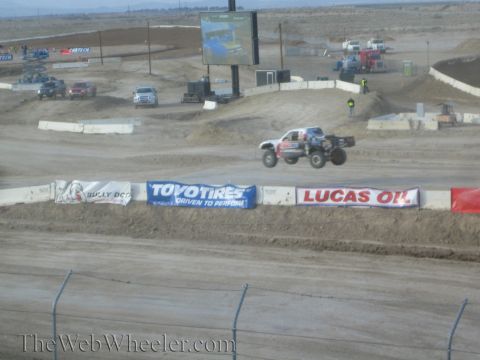 1990 Ford Bronco - white off-road race truck catches air over a jump on a desert short-course track past Toyo Tires and Lucas Oil banners.