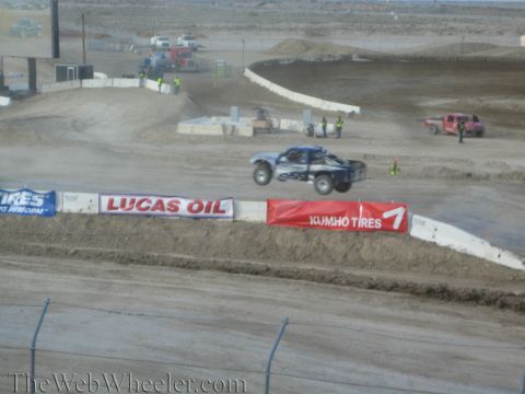 1990 Ford Bronco - blue off-road race truck launches over a berm on a desert short-course track past Lucas Oil and Kumho Tires banners.