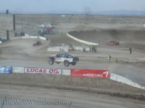 1990 Ford Bronco - off-road race truck races over a jump on a wide desert short-course track past Lucas Oil and Kumho Tires banners.