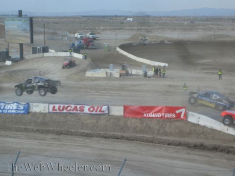 1990 Ford Bronco - Two off-road race trucks navigate jumps on a wide desert short-course track lined with Lucas Oil and Kumho Tires banners.