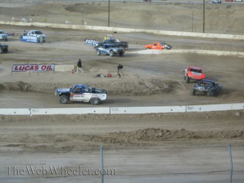 1990 Ford Bronco - Several off-road race trucks spread across a dusty dirt short-course track, with a blue truck leading past a Lucas Oil banner.