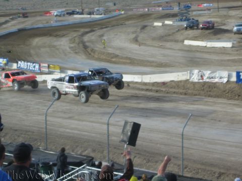 1990 Ford Bronco - Two off-road race trucks launch side by side off a jump on a dusty short-course track as the crowd cheers.