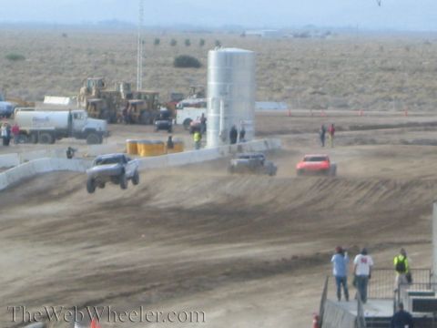 1990 Ford Bronco - Off-road race trucks run a dusty desert course near a large water tank and heavy equipment as people watch from the side.
