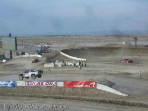 1990 Ford Bronco - off-road race truck launches over a jump on a wide desert short-course track past Lucas Oil and Kumho Tires banners.
