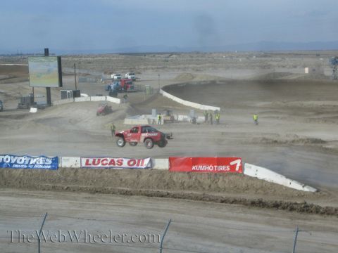 1990 Ford Bronco - red off-road race truck catches air over a jump on a wide desert short-course track past Lucas Oil and Kumho Tires banners.