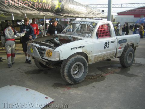 1990 Ford Bronco - muddy white number 83 off-road race truck with Toyo Tires graphics sits in a paddock as crew members stand nearby.