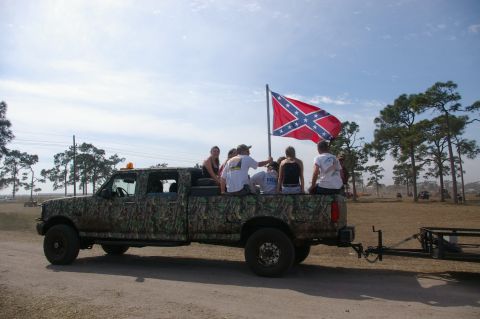 1974 Ford F-100 - A camouflaged pickup truck carries passengers and a confederate flag, with a trailer attached.