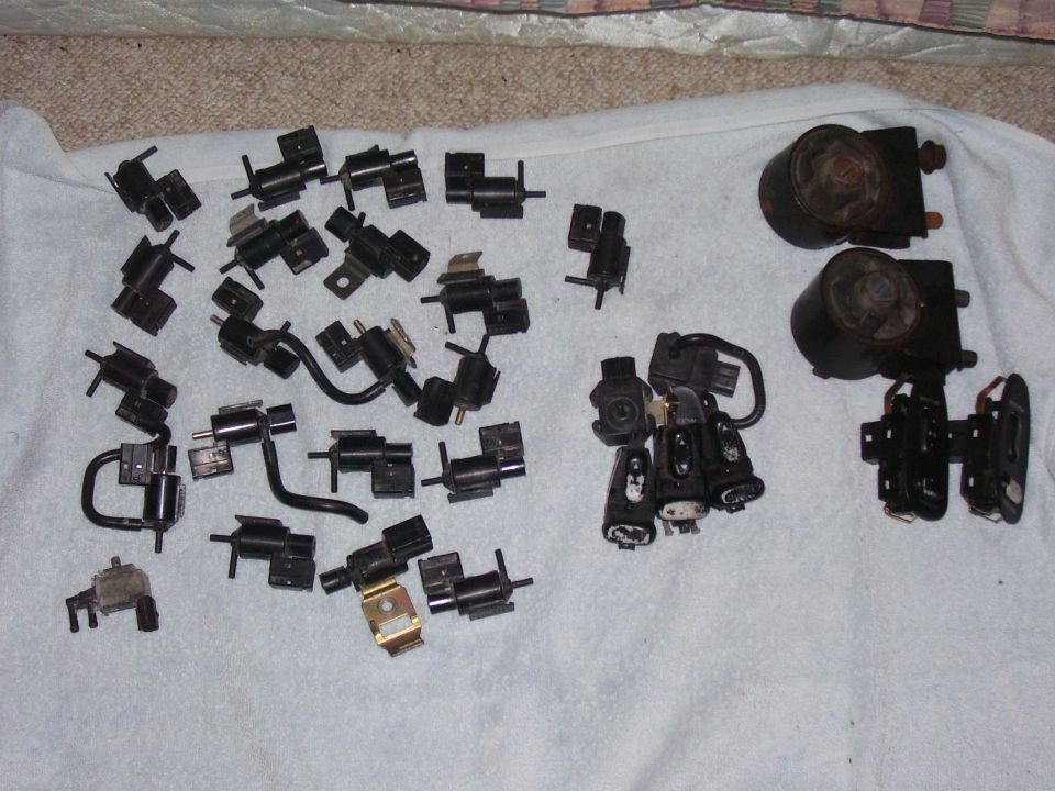 Pile of various black plastic and metal automotive vacuum solenoids and actuators laid out on a white cloth