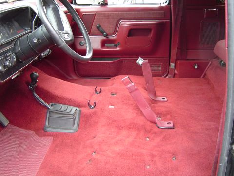 1989 Ford Bronco - The interior of a red vehicle shows a bare floor with a manual transmission and seatbelt anchors, suggesting recent work or customization.