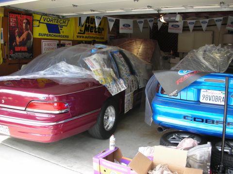 1992 Ford Crown Victoria - Two cars, a red sedan and a blue Mustang Cobra, are parked in a garage with various automotive banners on the wall.