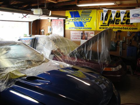 1992 Ford Crown Victoria - Two vehicles are covered in plastic sheeting inside a garage, with various automotive banners on the wall.