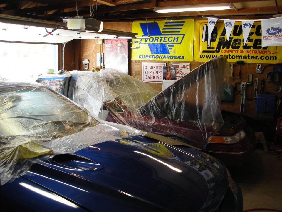 Two cars covered in plastic in a garage. One is blue with a hood scoop, the other is maroon