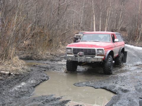 1990 Ford F-250 - This red Ford Bronco navigates a muddy trail with trees on either side, while another vehicle follows.