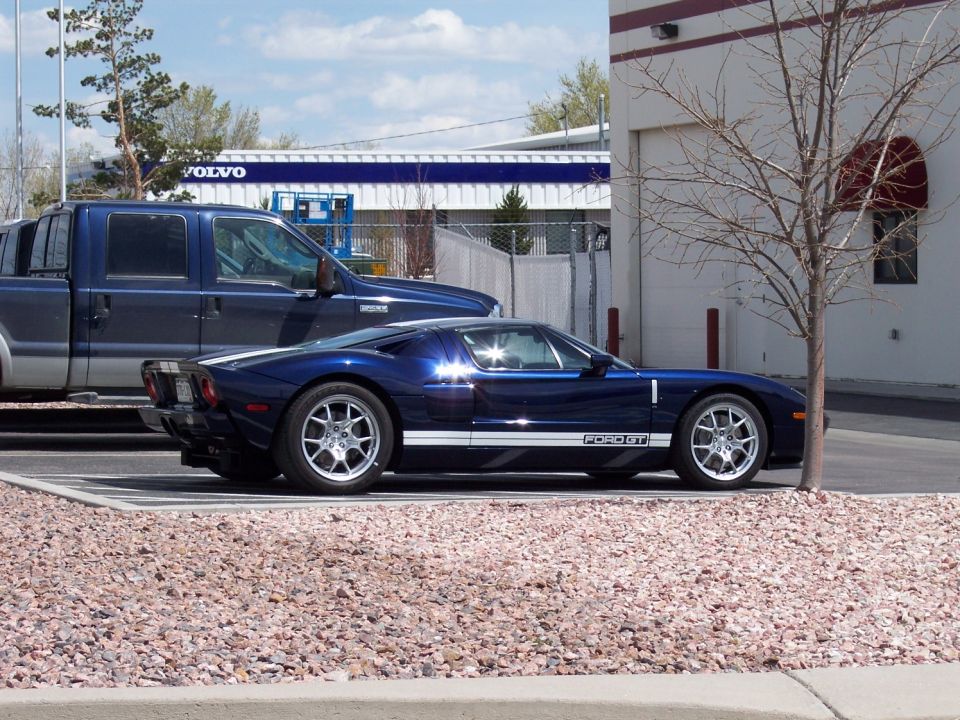 Blue Ford GT sports car with white racing stripes parked next to a blue pickup truck. "FORD GT" text visible on the side