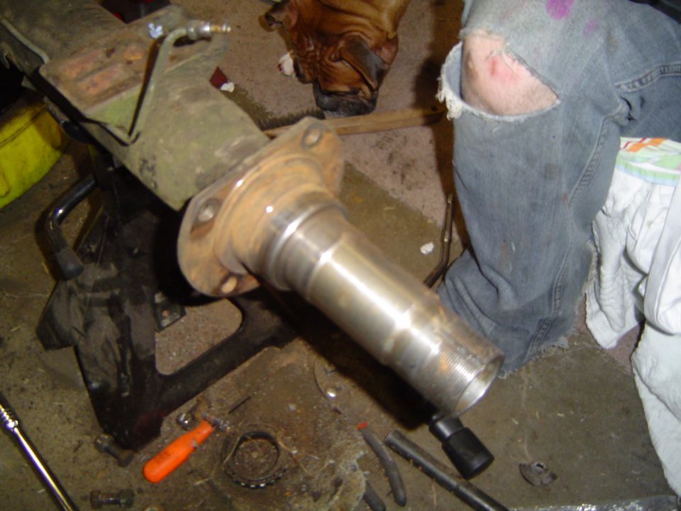 Close-up of a metal axle component on a jack stand, with a person's ripped jeans and a dog in the background