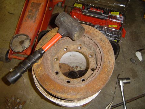 1978 Ford Bronco - A rusty brake rotor sits on a bucket, with a mallet resting on it, alongside a floor jack and a red toolbox.