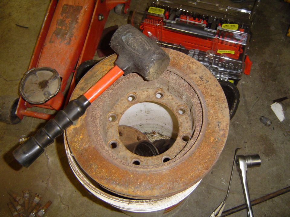 Rusty 8-lug vented brake rotor on shop floor with sledgehammer and floor jack