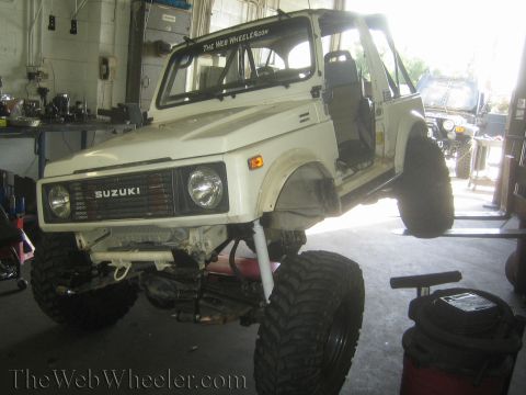 1987 Suzuki Samurai - White 1987 Suzuki Samurai is being worked on, lifted by a forklift in a workshop. A second vehicle is in the background.