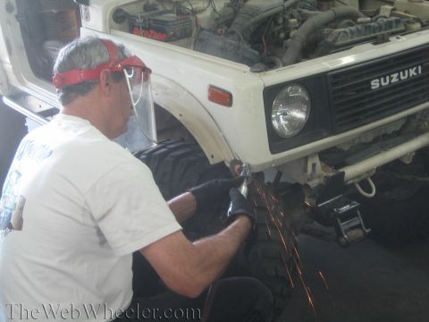 1987 Suzuki Samurai - A man grinds on the front fender of a white 1987 Suzuki Samurai, with the hood open revealing the engine.