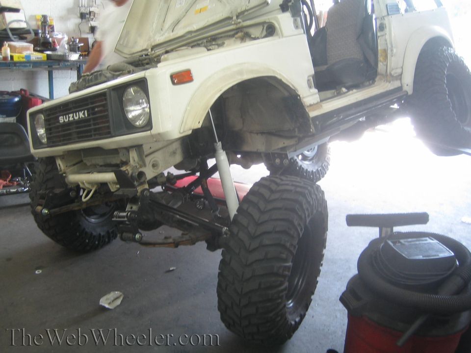 1987 Suzuki Samurai lifted with large off-road tires, hood open, in a garage