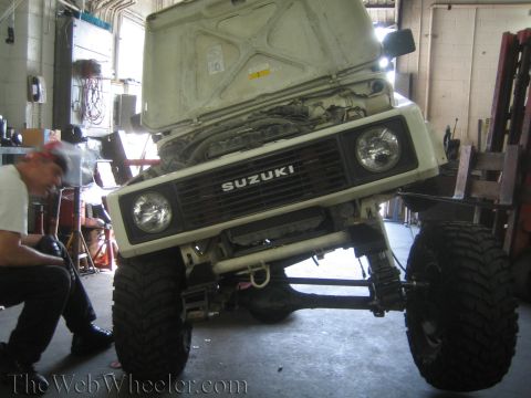 1987 Suzuki Samurai - Front view of a 1987 Suzuki Samurai in a workshop with its hood raised, revealing the engine bay.