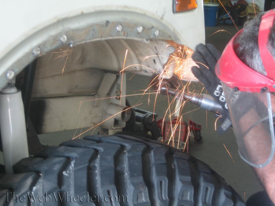Person wearing safety gear grinds metal on a vehicle's wheel well near a large tire
