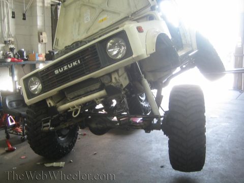 1987 Suzuki Samurai - A white 1987 Suzuki Samurai is shown lifted with large off-road tires in a garage setting.