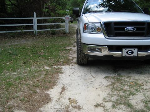 2004 Ford F-150 - A white Ford F-150 is parked on a dirt driveway next to a white fence, with a 'G' emblem on the front license plate.