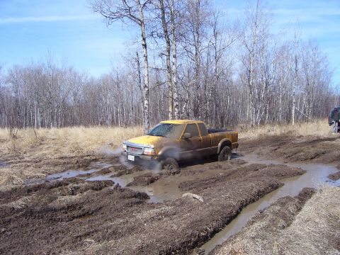 1996 Ford Bronco - A yellow pickup truck navigates a muddy off-road trail with another vehicle visible in the background.