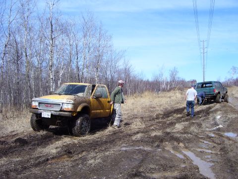 1996 Ford Bronco - Two trucks navigate a muddy off-road course under a bright blue sky.