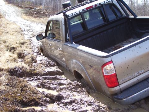 1996 Ford Bronco - A silver Toyota Tundra is stuck in thick mud on an unpaved road, with its rear passenger side wheel submerged.